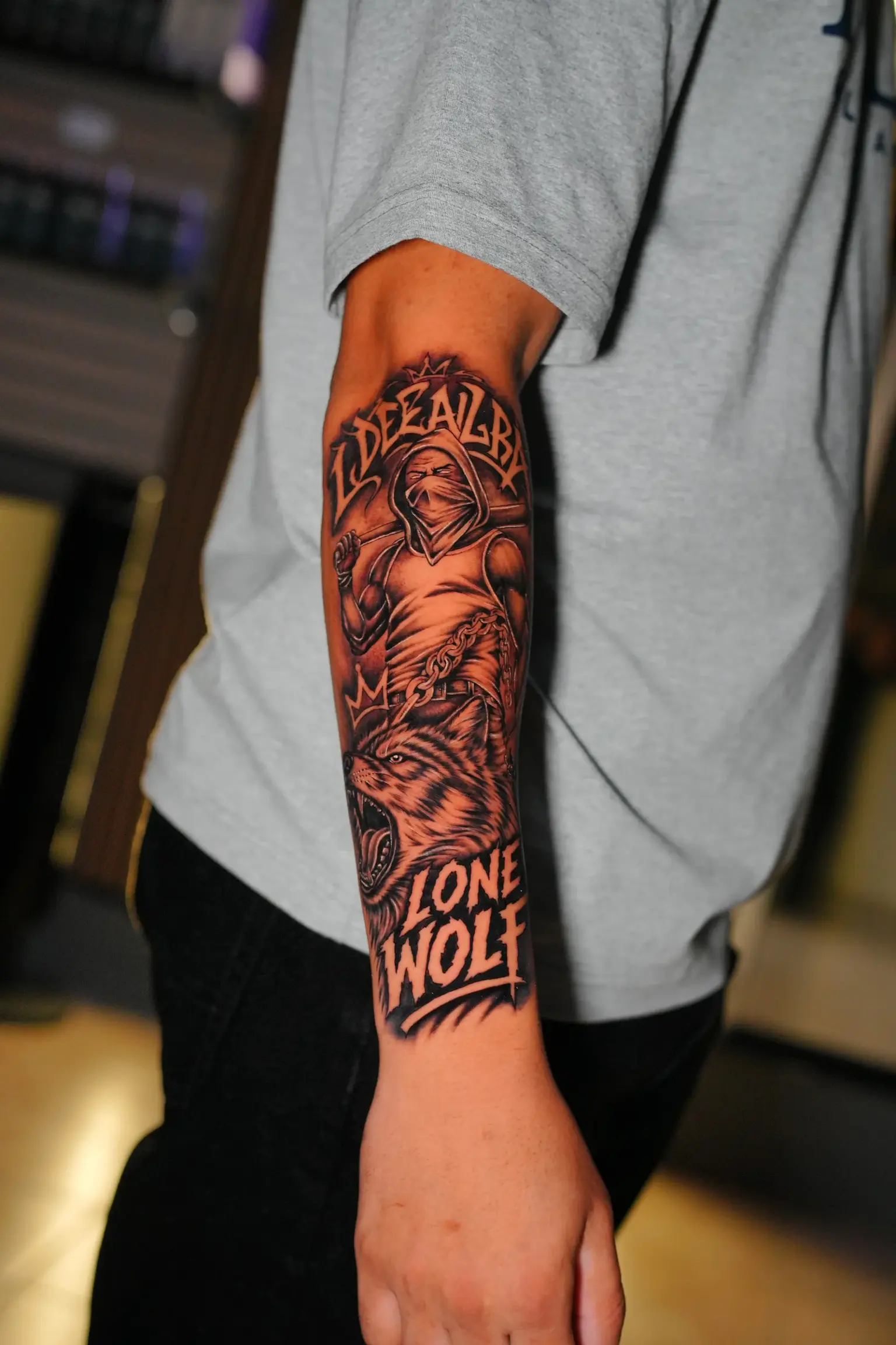 Black and grey forearm tattoo of snarling lone wolf, masked streetwear figure, and crown lettering
