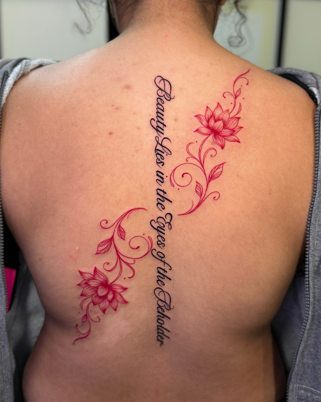 Elegant script tattoo reading Beauty Lies in the Eyes of the Beholder running down the spine, framed by two large red lotus flowers and vines